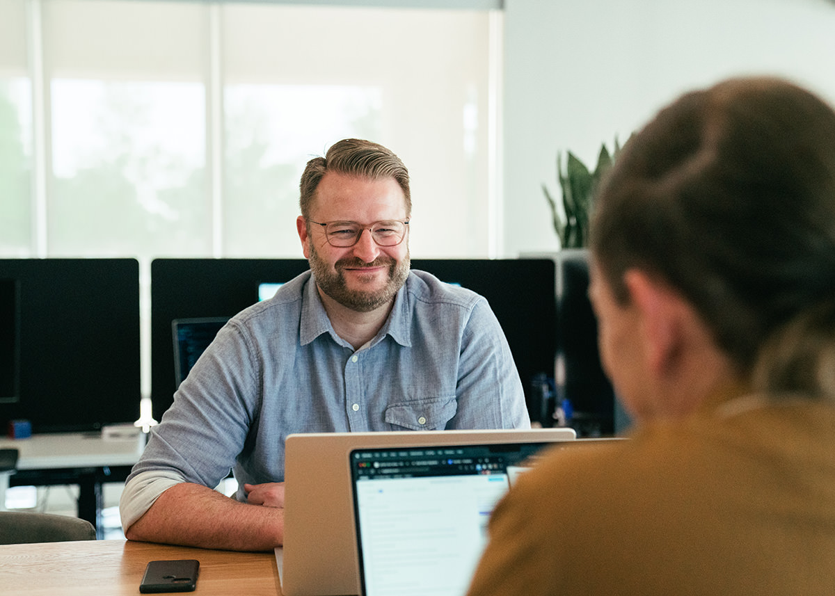 A ruggedly handsome man, smiling while talking to another person across the table.