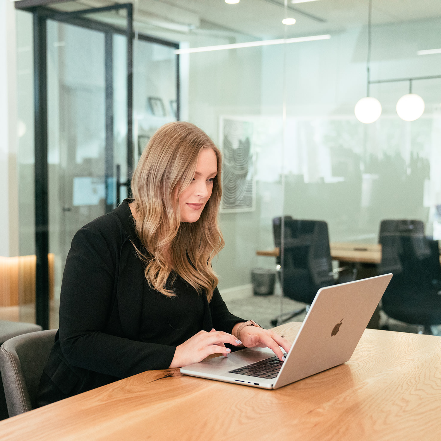 A woman smiling while sitting at a table and working on her laptop in a large open office area.