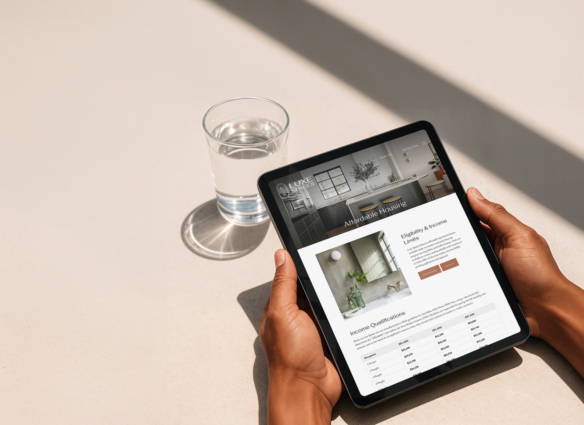 A person holding a tablet with a property's affordable housing page and a glass of water in the background.