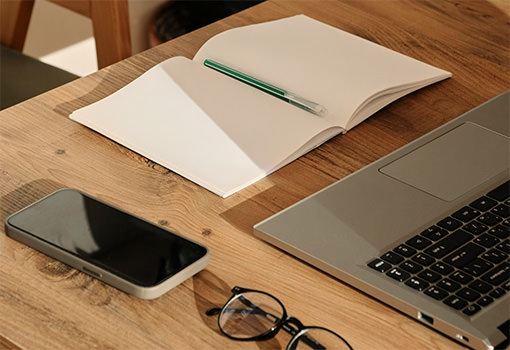 A desk with a phone, notebook, pen, laptop and glasses in the sunlight.