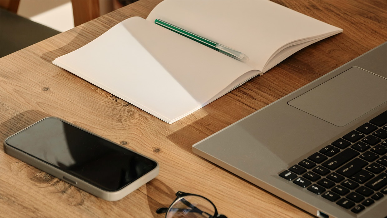 A desk with a phone, notebook, pen, laptop and glasses in the sunlight.