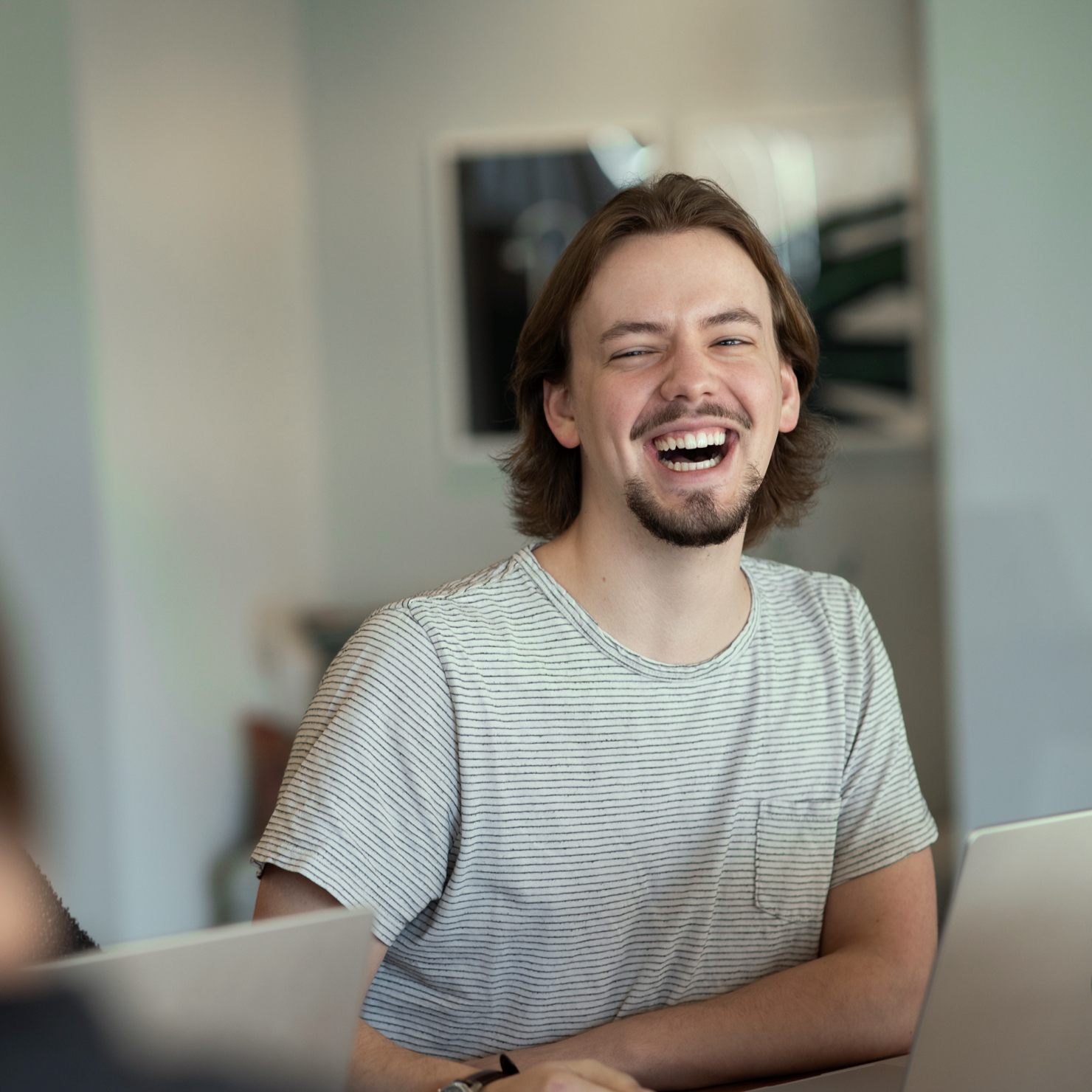 A  young man sitting and laughing in an open office area.